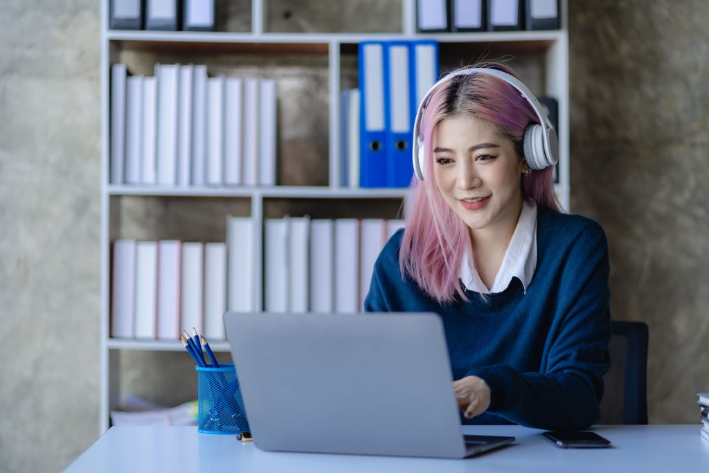 Asian female college student wearing headphones studying online with laptop computer surfing internet meeting Studying online, smiling happily at home.